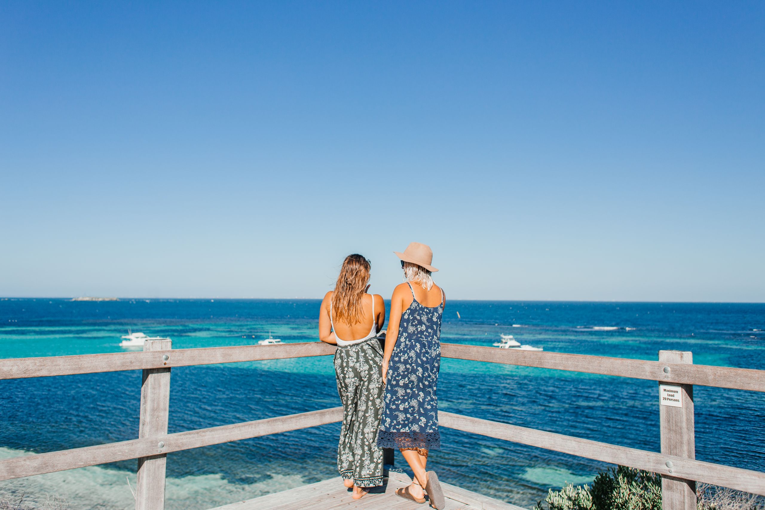 Two women stand on a viewing platform overlooking the ocean, enjoying a sunny day.